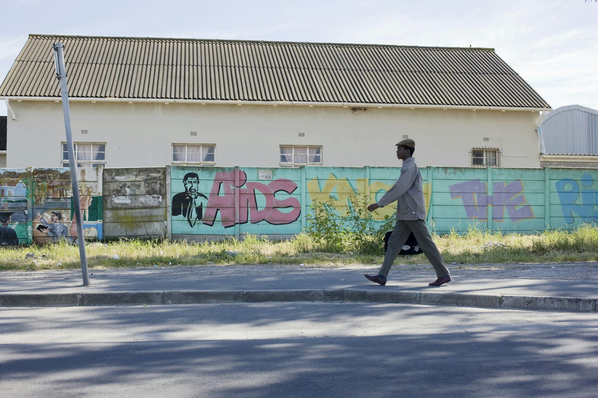 A wide shot of a wall with graffitti and a young African man walking resolutely in work clothes.