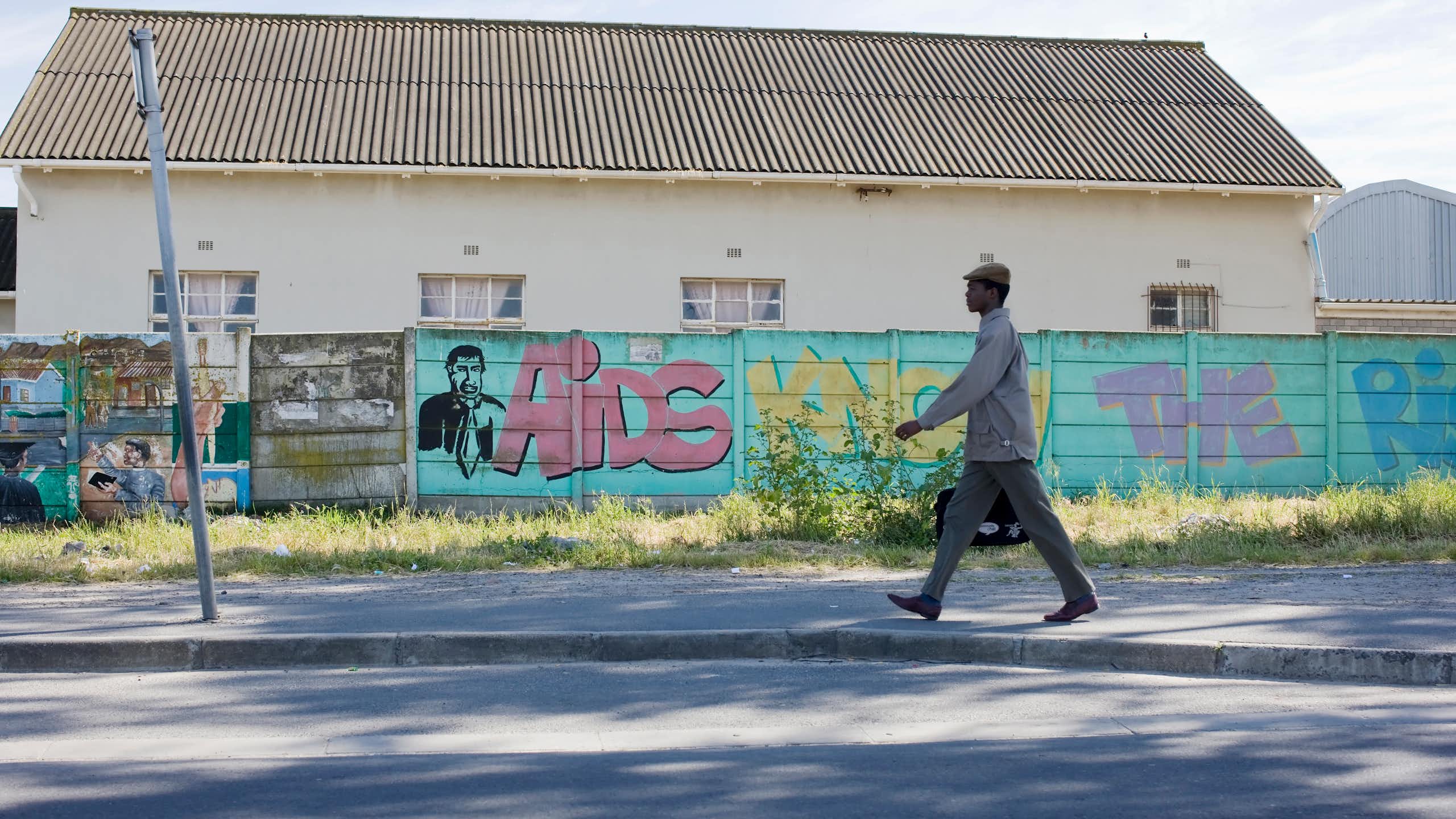 A wide shot of a wall with graffitti and a young African man walking resolutely in work clothes.