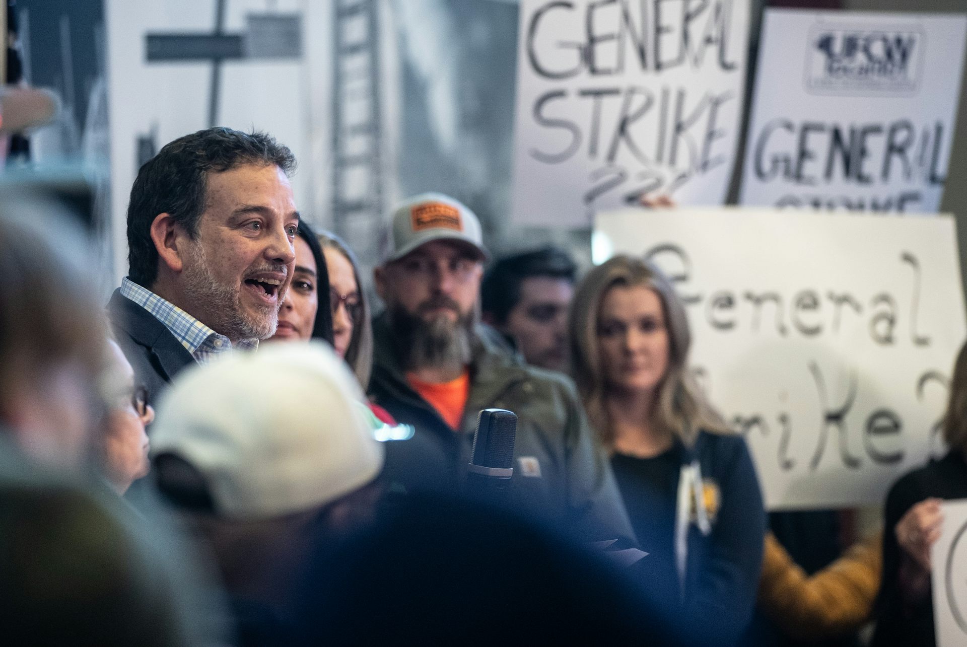 A man with dark hair speaks to a crowd of people, some carrying signs that read General Strike.