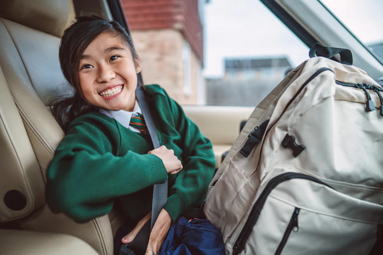 A girl in school uniform smiles as she undoes her seatbelt in a car.