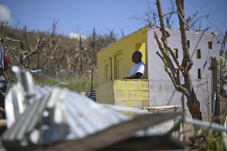 A man stands at the door of a home that has lost its roof. Its metal roof is crumpled on the ground, and it is surrounded by trees that were stripped of all their leaves by hurricane winds.