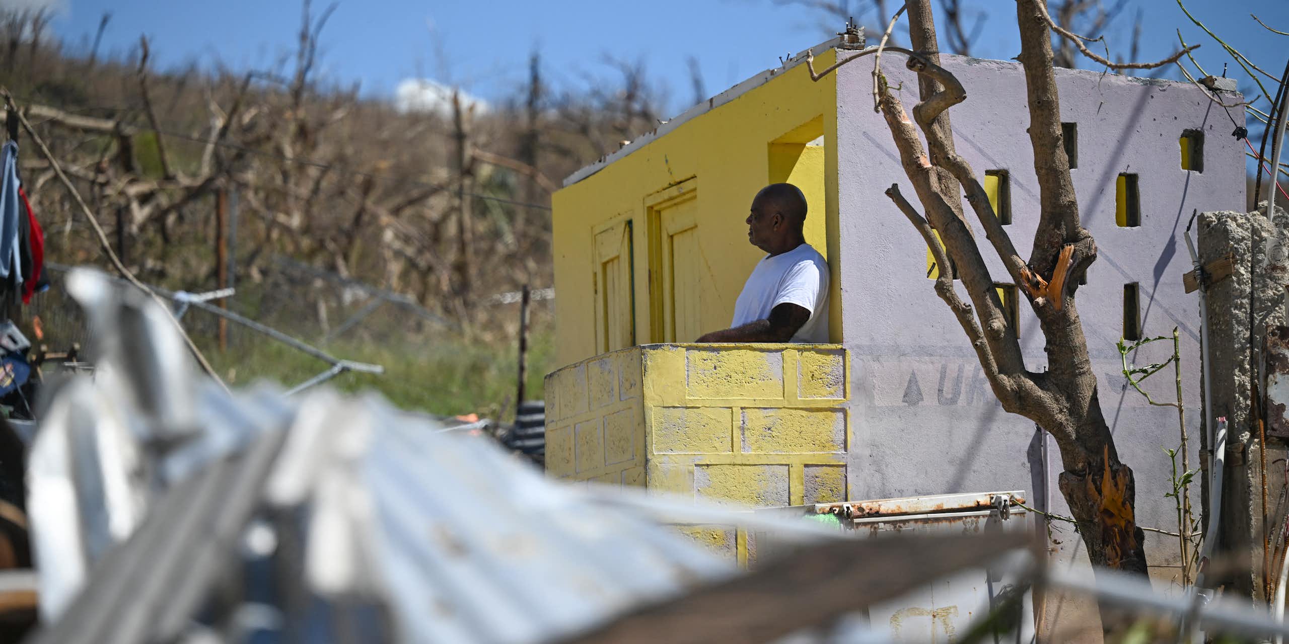 A man stands at the door of a home that has lost its roof. Its metal roof is crumpled on the ground, and it is surrounded by trees that were stripped of all their leaves by hurricane winds.