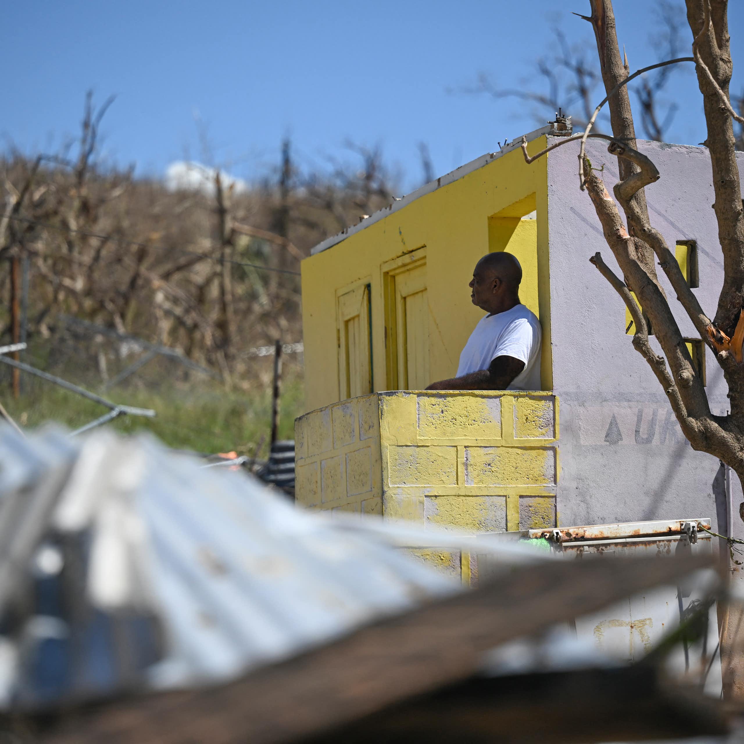 A man stands at the door of a home that has lost its roof. Its metal roof is crumpled on the ground, and it is surrounded by trees that were stripped of all their leaves by hurricane winds.
