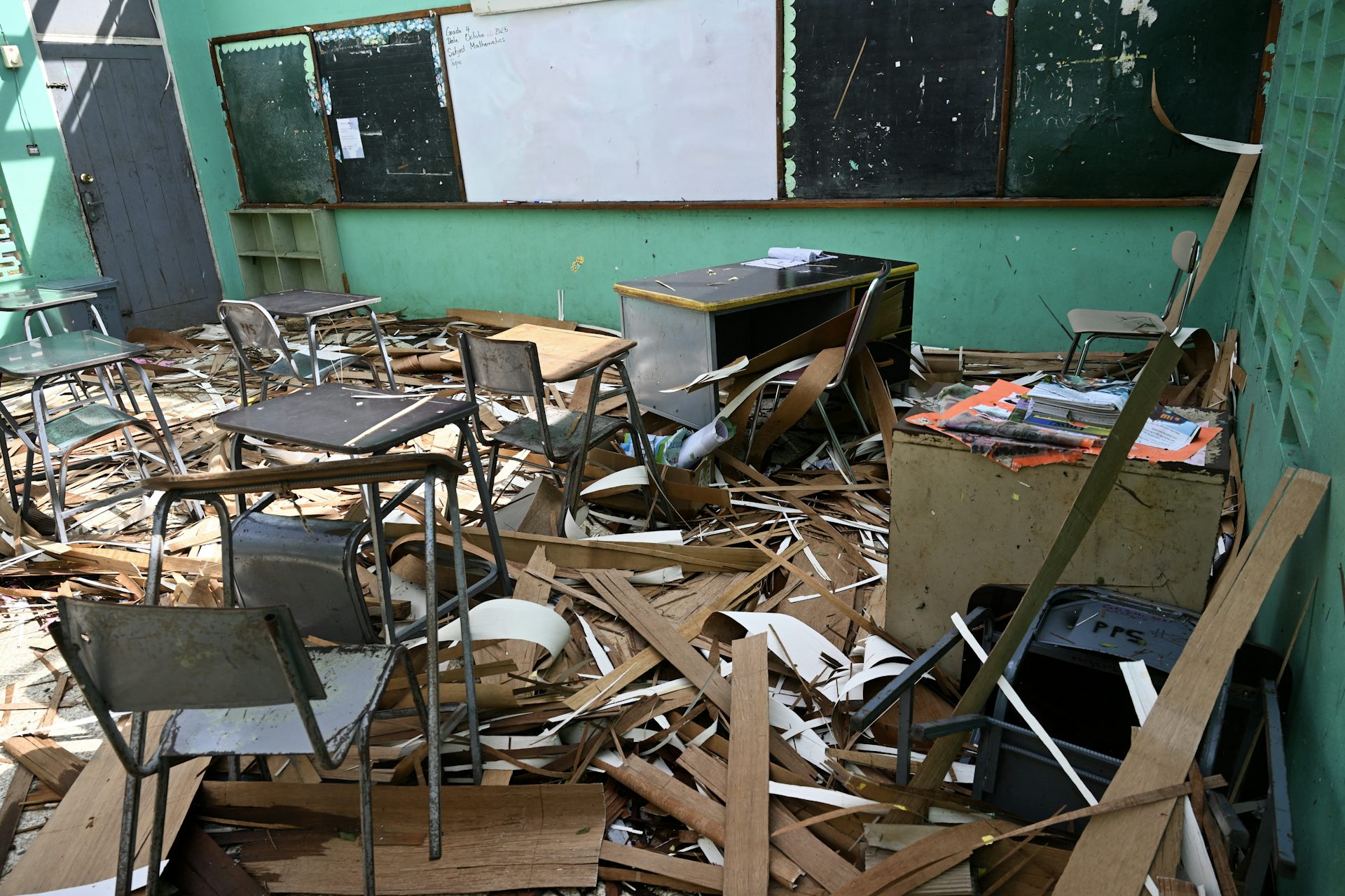 The interior of a school that has been torn apart by hurricane winds. Desks and debris are scattered and light shines through the rafters