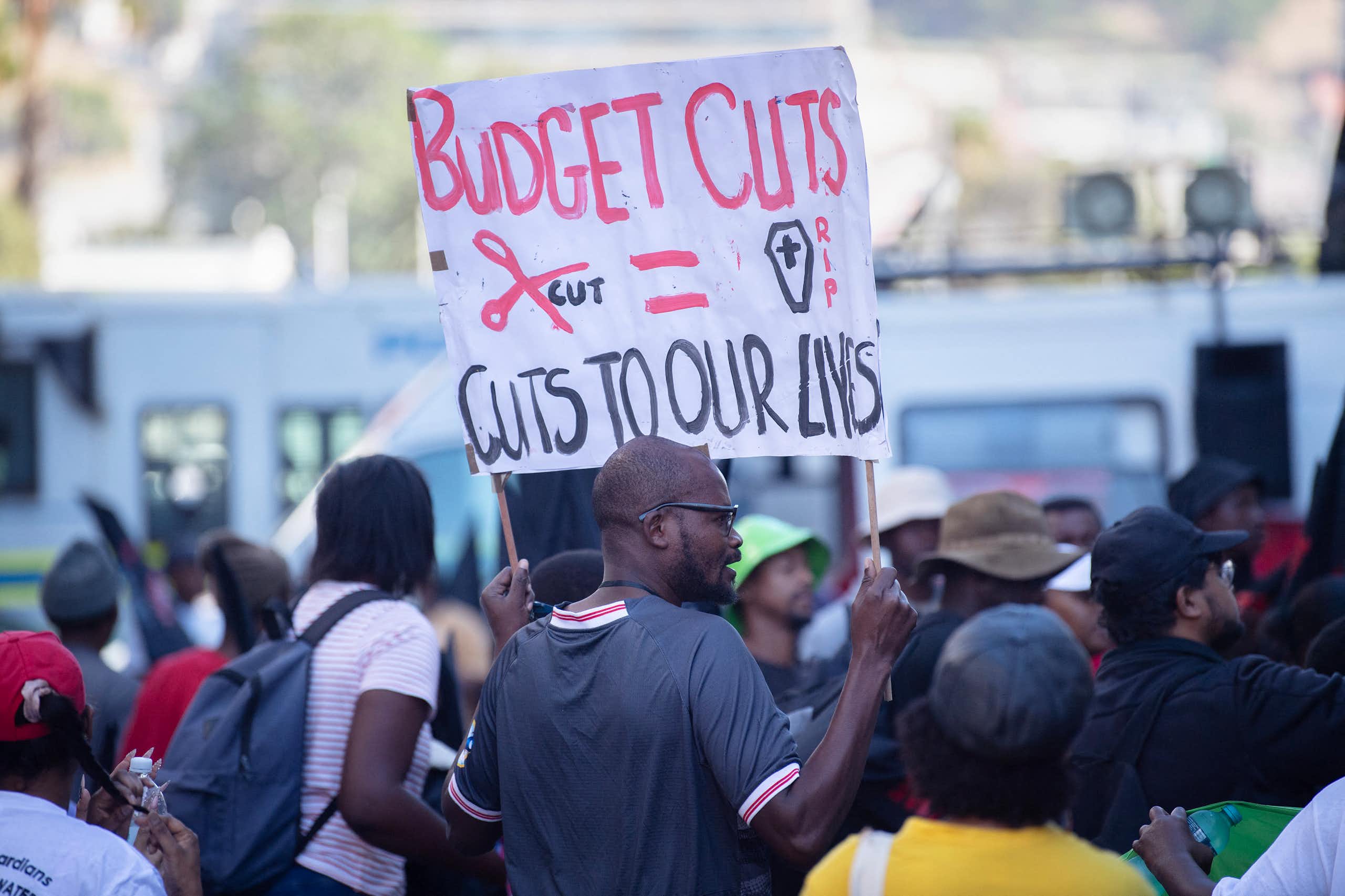 Group of people, one holding up a banner saying 'Budget cuts = cuts to our lives"