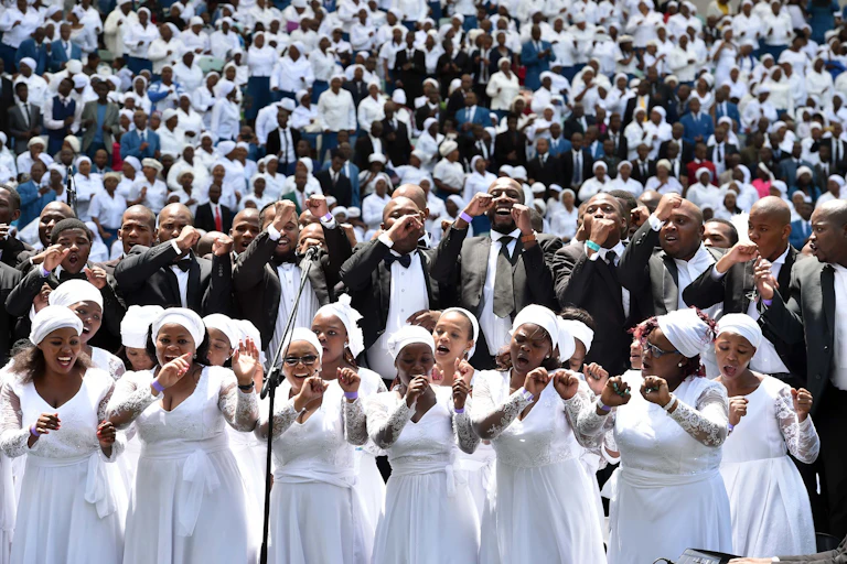 A joyful church choir of women in white dresses with a choir of men in suits above them and a stand packed with people behind them. They sing, making arm movements.
