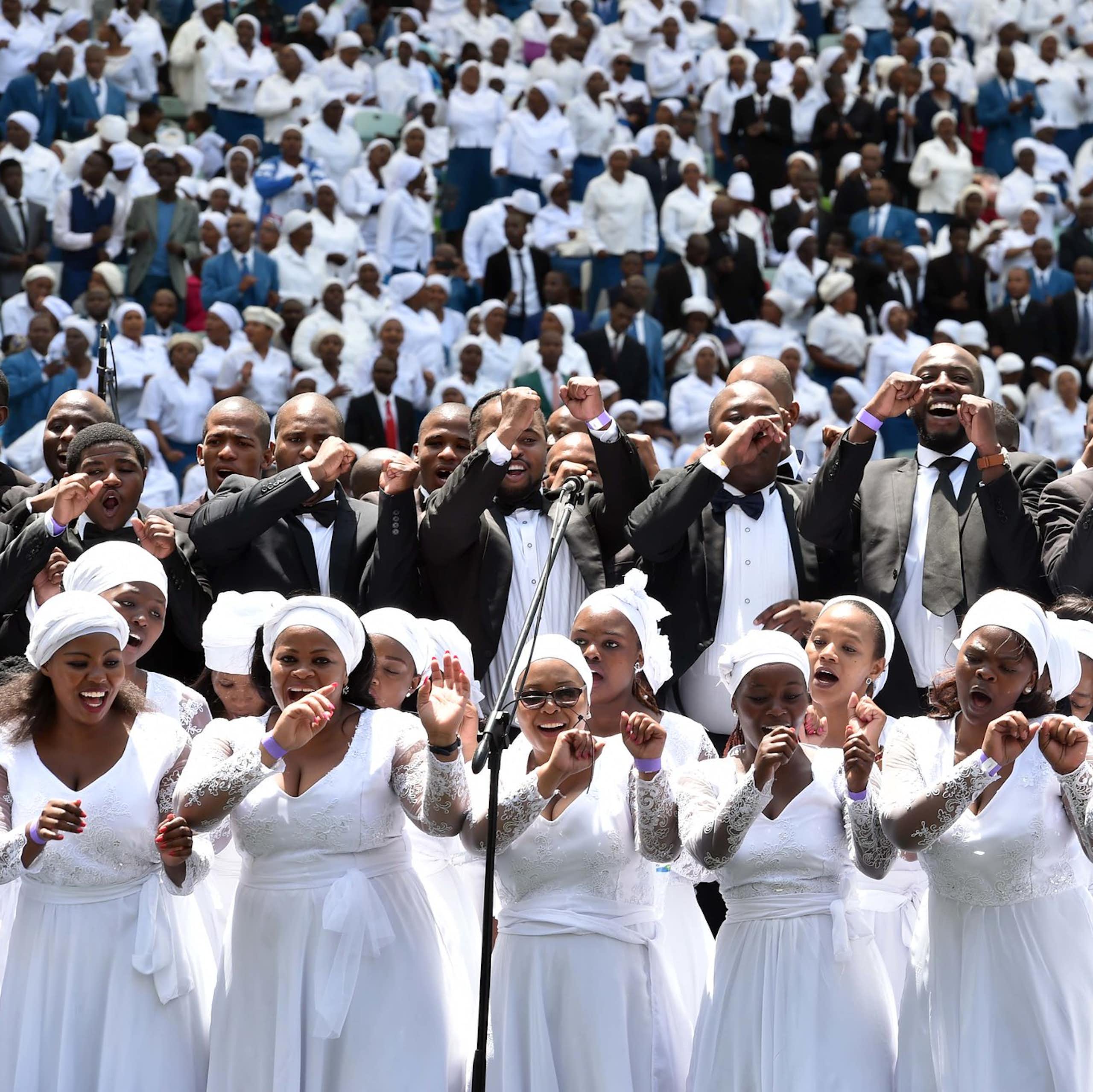 A joyful church choir of women in white dresses with a choir of men in suits above them and a stand packed with people behind them. They sing, making arm movements.