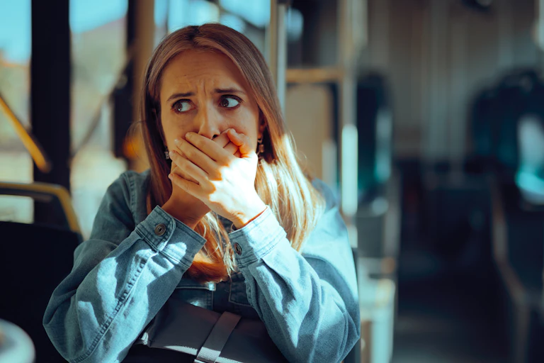 A woman sitting on a bus covers her mouth while looking fearful.