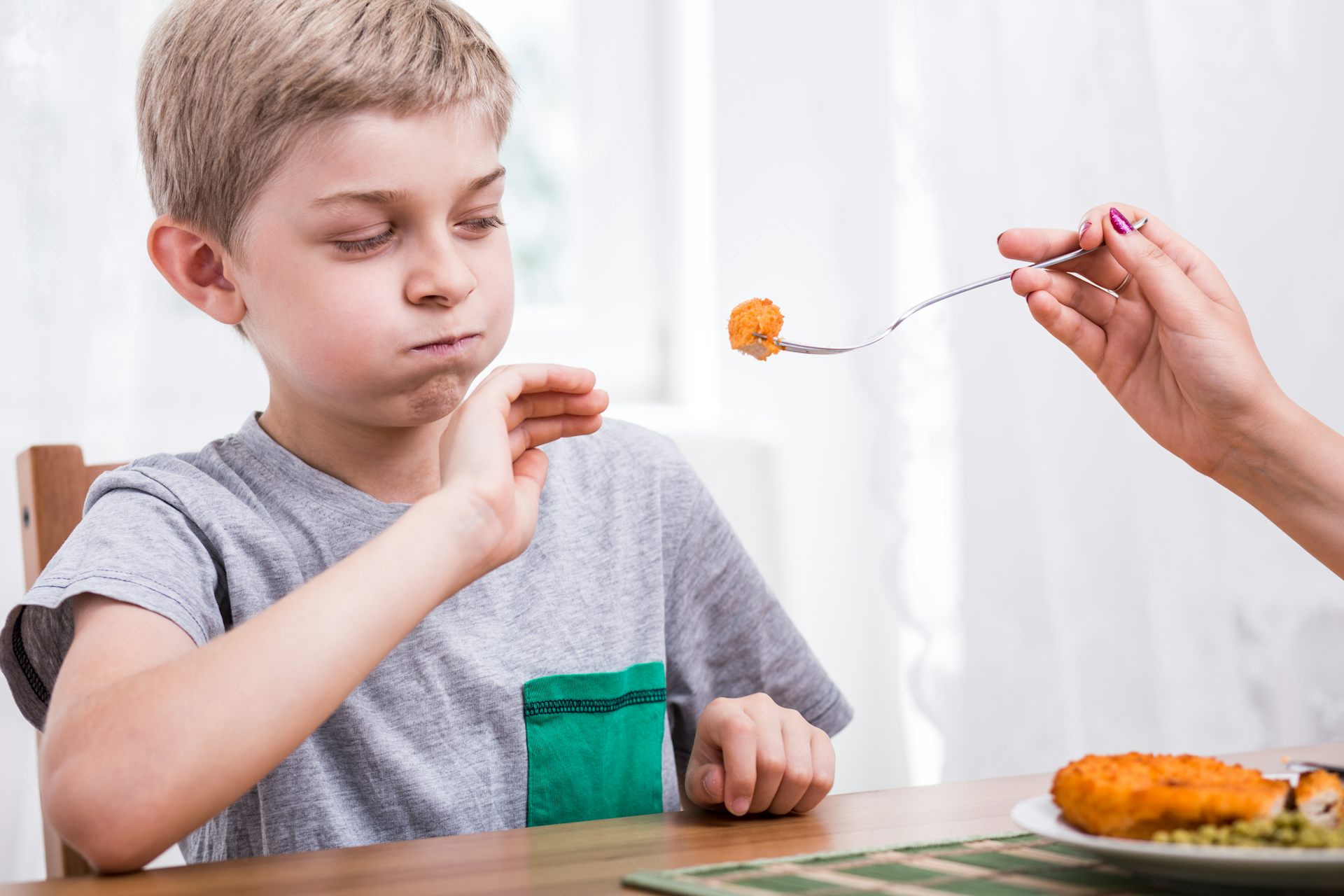 A young boy refuses the breaded meat which is being offered to him on a fork by another person.