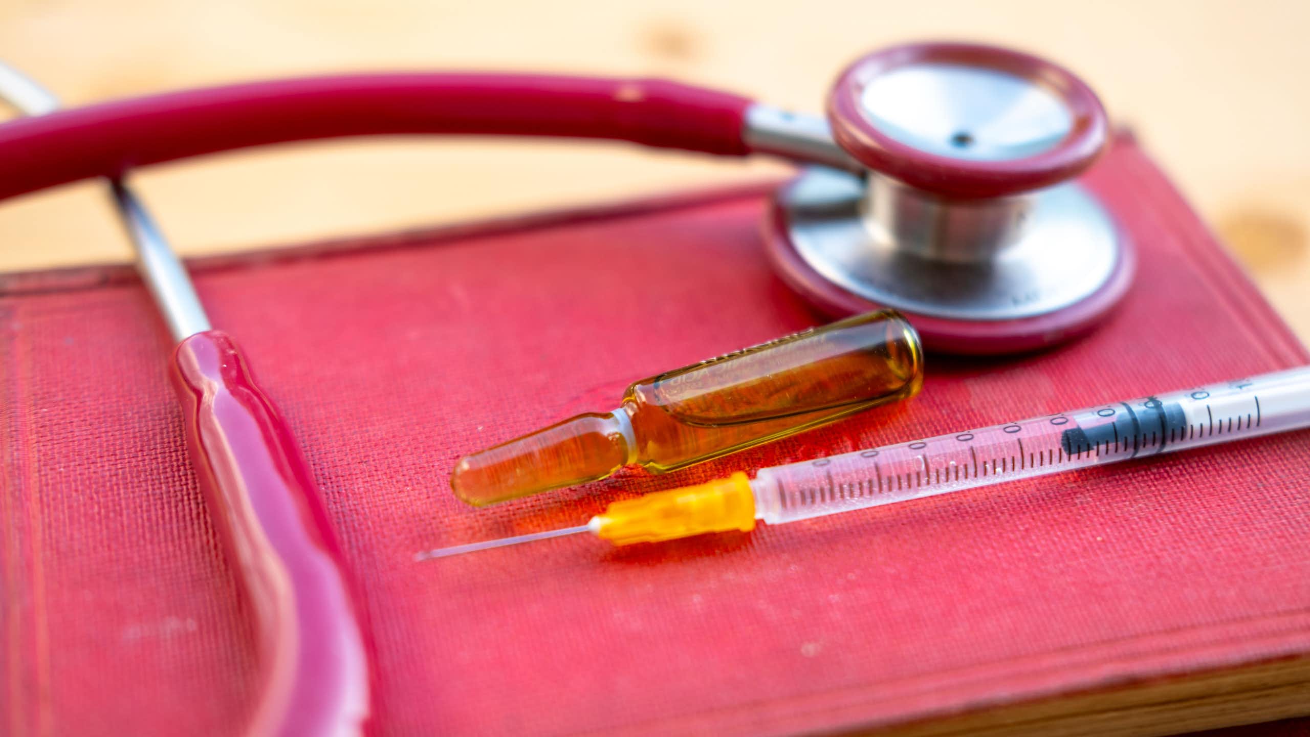 Une seringue, un stéthoscope et une fiole contenant du matériel d’injection sont posés sur un livre rouge représentant la loi.
