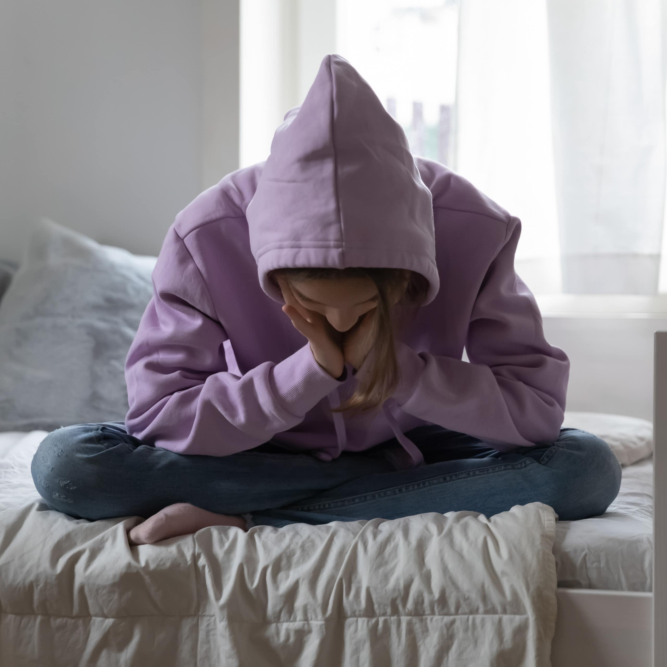 Une jeune fiche en sweatshirt à capuche, tête baissée, se tient le visage entre les mains, l'air déprimé.