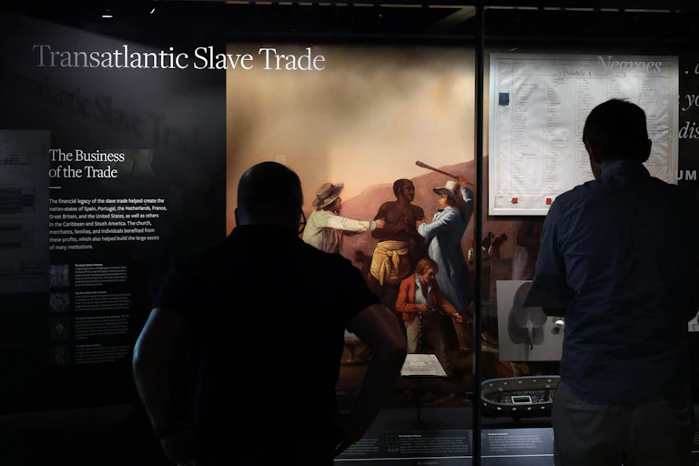 silhouettes of two visitors look at an image showing two white people forcibly handling a black slave, in an exhibition about slavery in The United States of America at the National Museum of African American History and Culture on August 28, 2025 in Washington, DC.