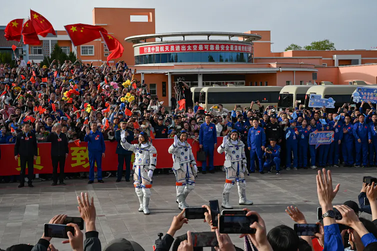 Three astronauts walking down a street lined with crowds in stands waving Chinese flags.