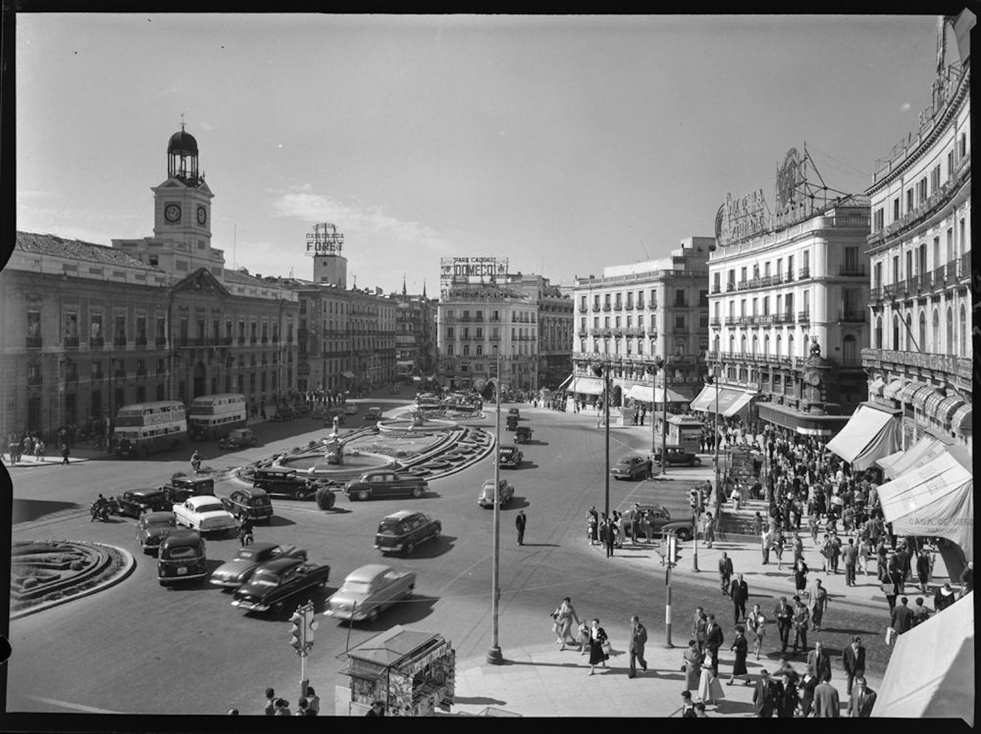 Vista de la Puerta del Sol con el emblemático reloj a la izquierda, tráfico circulando por la plaza y transeúntes en las aceras bajo los toldos de los comercios. Carteles luminosos publicitarios en las azoteas de los edificios
