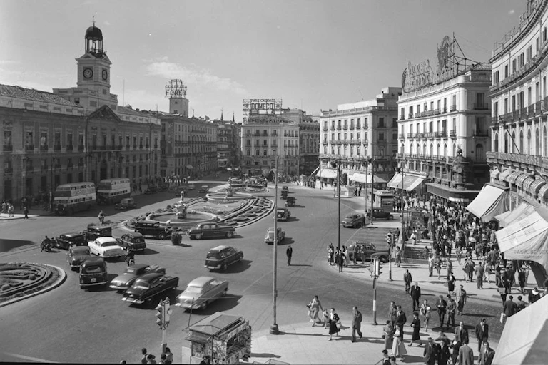 Vista de la Puerta del Sol con el emblemático reloj a la izquierda, tráfico circulando por la plaza y transeúntes en las aceras bajo los toldos de los comercios. Carteles luminosos publicitarios en las azoteas de los edificios
