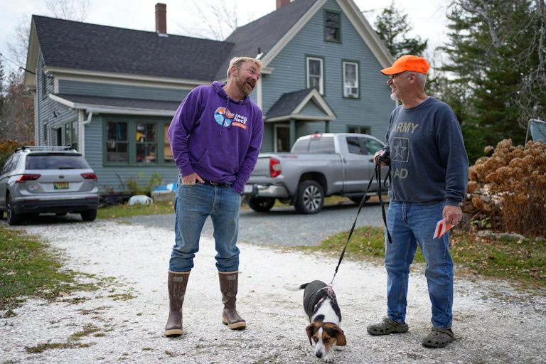 Two men and a dog on a leash in a driveway in front of a home.