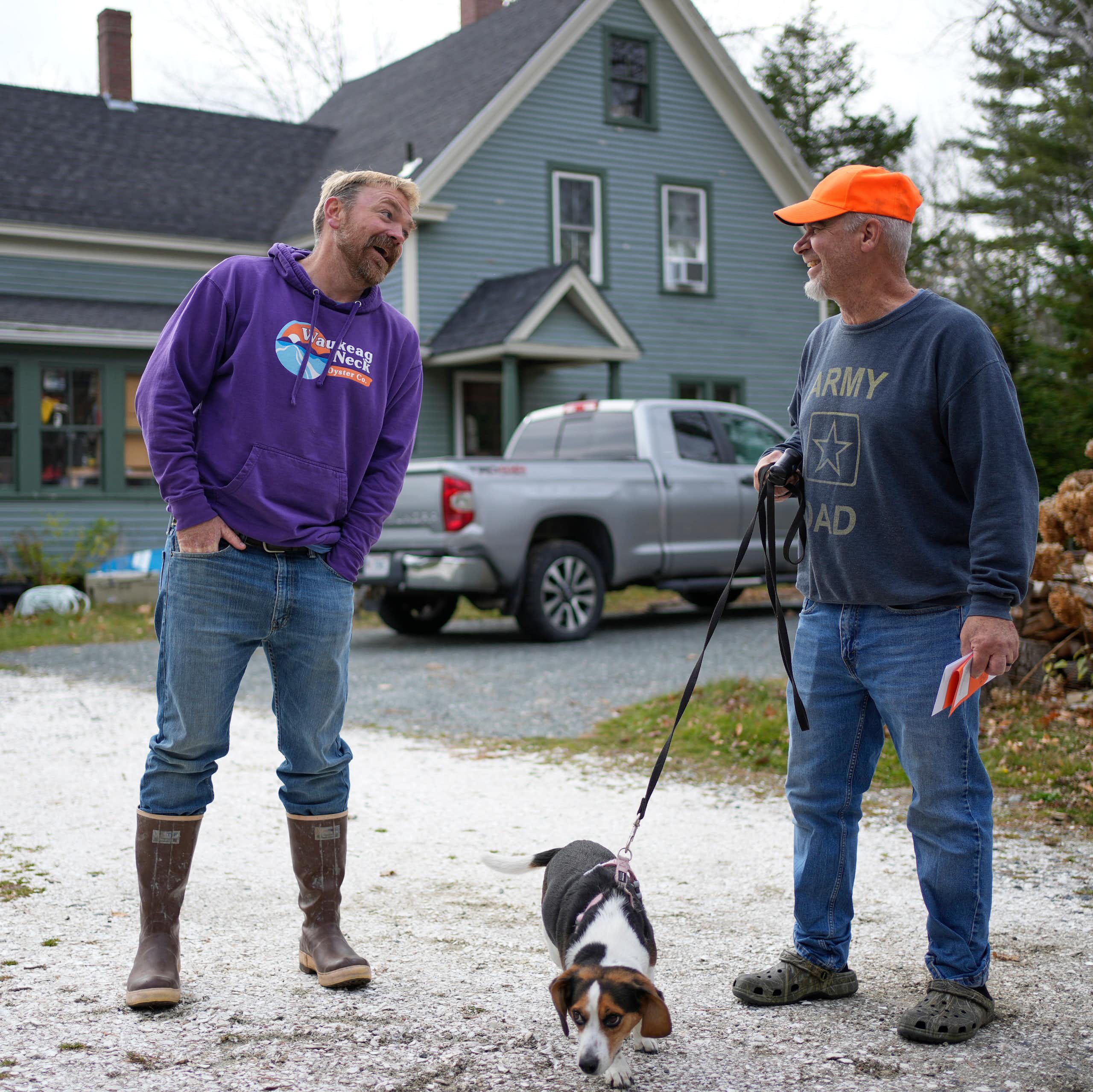 Two men and a dog on a leash in a driveway in front of a home.