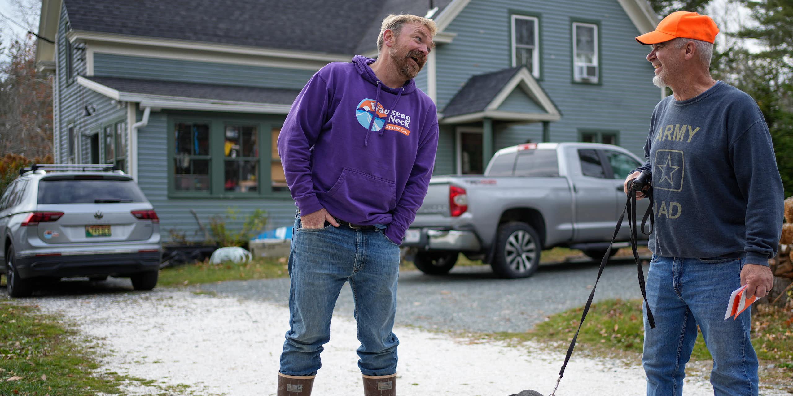 Two men and a dog on a leash in a driveway in front of a home.