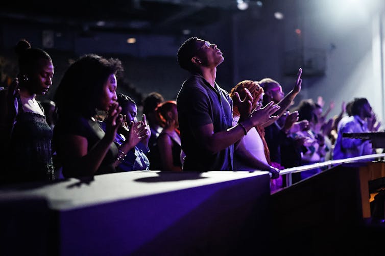 Rows of young people, most of whom are Black, stand and sing in a large darkened auditorium.