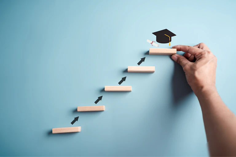 A small ladder leads to a person holding up one of the ladder footrests, with a black graduation cap on top of it.