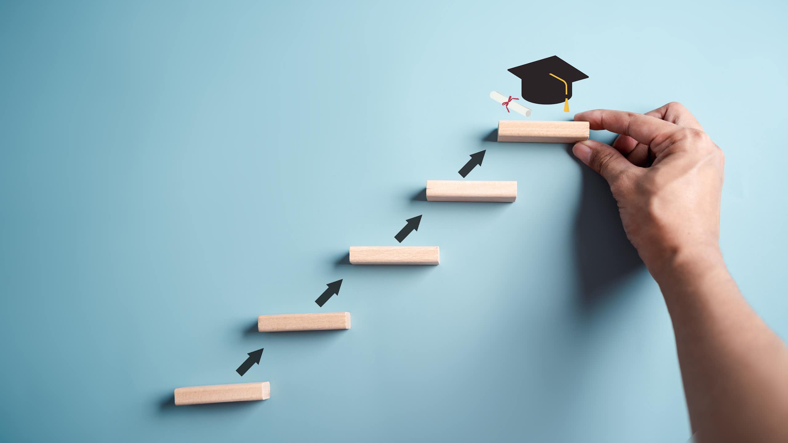 A small ladder leads to a person holding up one of the ladder footrests, with a black graduation cap on top of it.