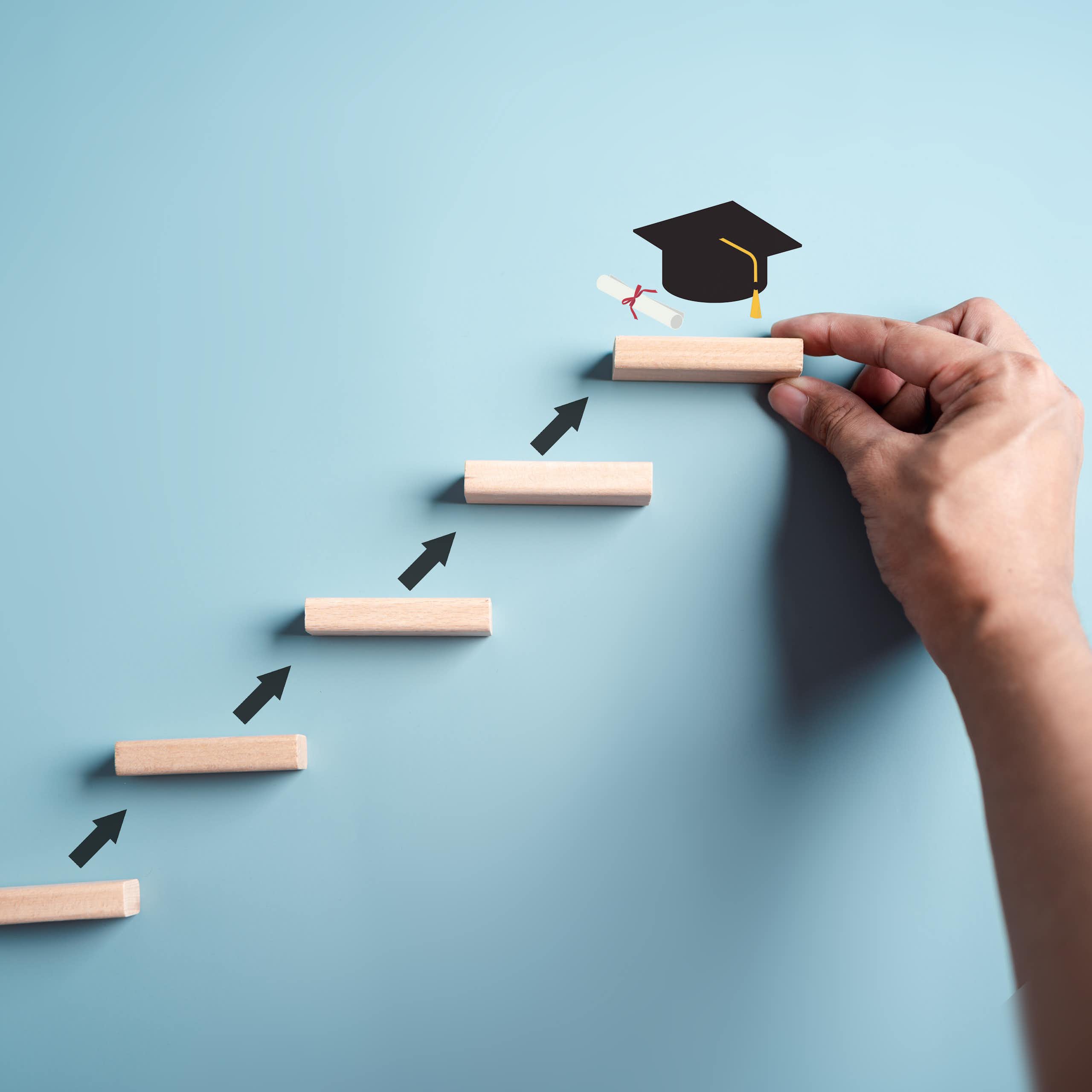 A small ladder leads to a person holding up one of the ladder footrests, with a black graduation cap on top of it.