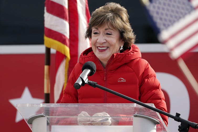 A woman in a red parka speaking into a microphone at a lectern, in front of an American flag.