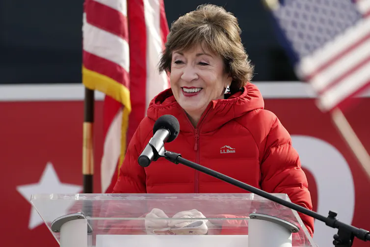 A woman in a red parka speaking into a microphone at a lectern, in front of an American flag.