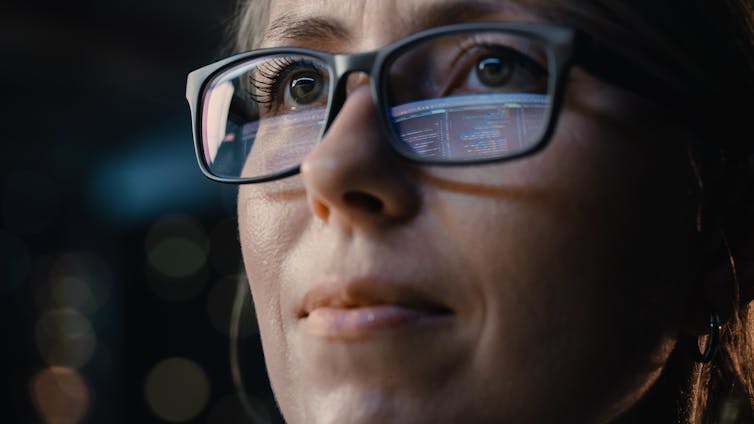 a woman looks pensive with computer code reflected in her glasses.