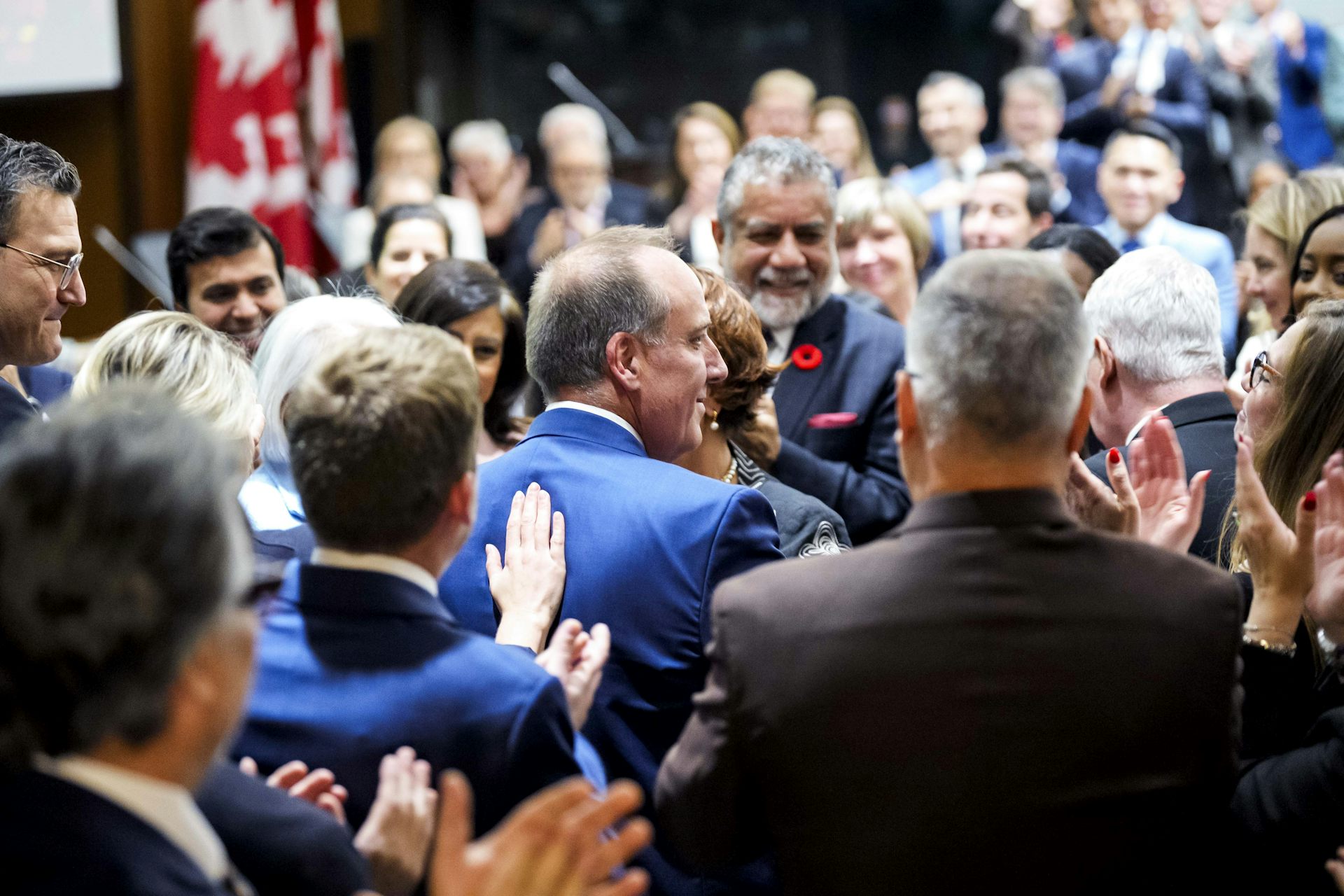 A man in a blue suit with thinning hair smiles as people pat him on the back.