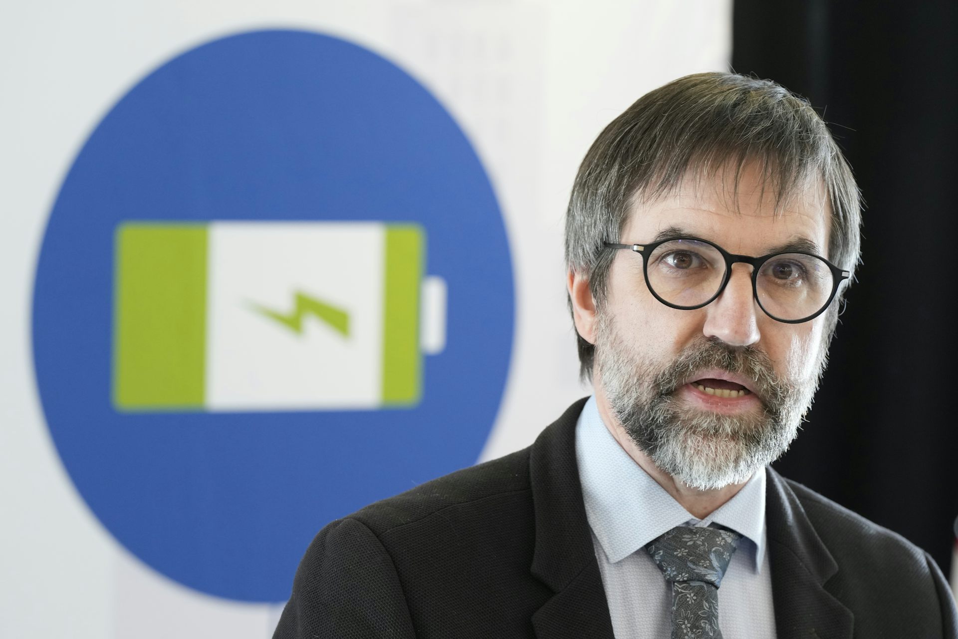 A man with a grey beard and dark grey hair wearing glasses speaks in front of a battery illustration.