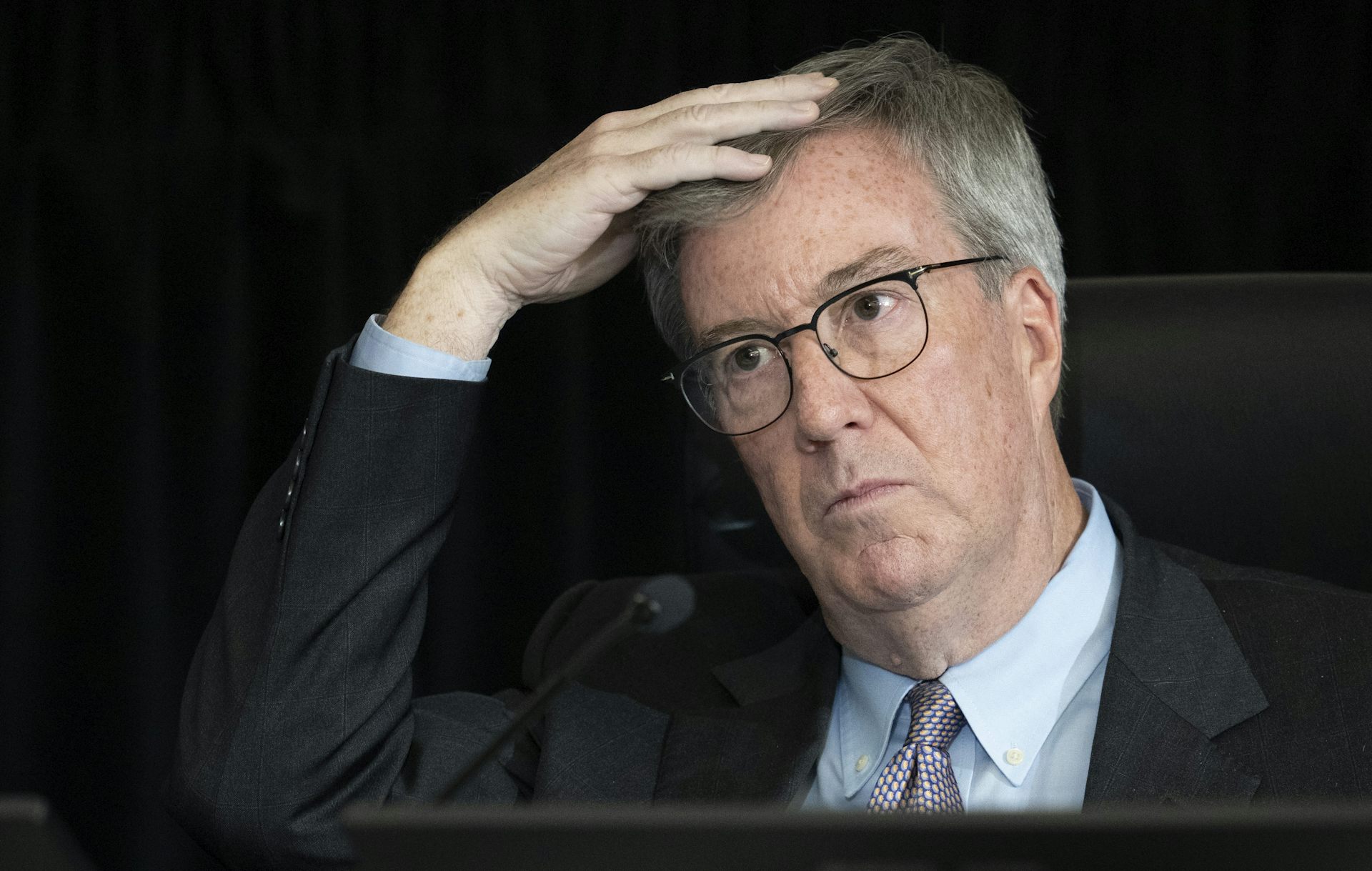 A man with grey hair and glasses with one hand on his head.