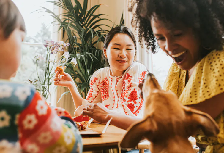 Trois personnes assises à table en train de manger et de sourire à un chien qui demande un morceau