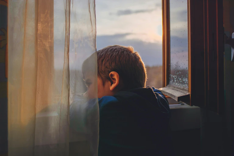 Glum little boy, seen through gauze curtain, stares out an open window.