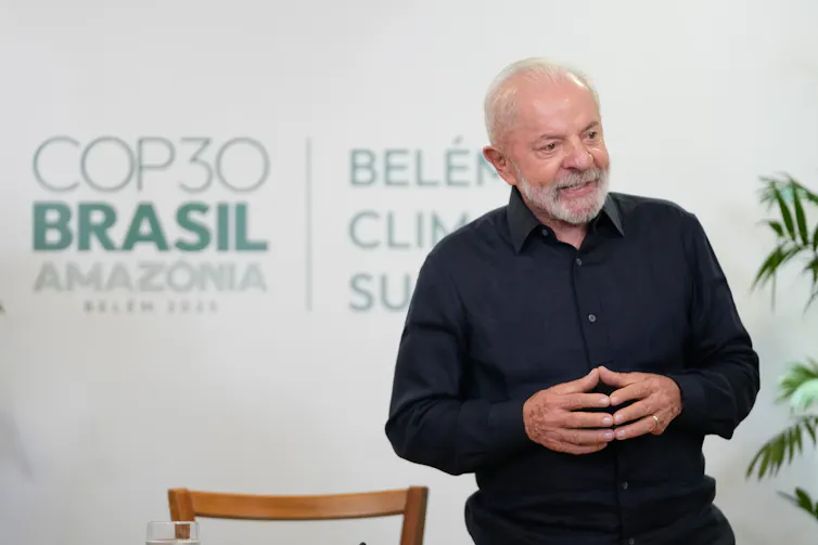 an older man with white hair and beard wearing black clothes stands in front of a sign reading COP30 Brasil