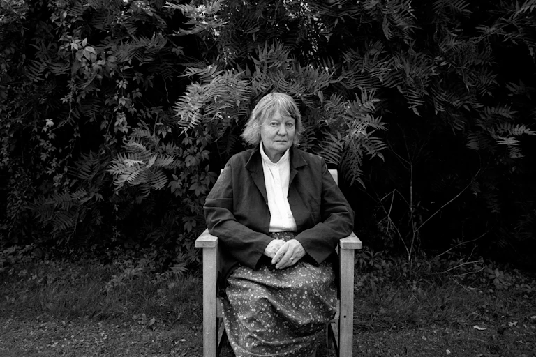 A black and white image of an older woman sitting on a chair in a garden looking sombre.