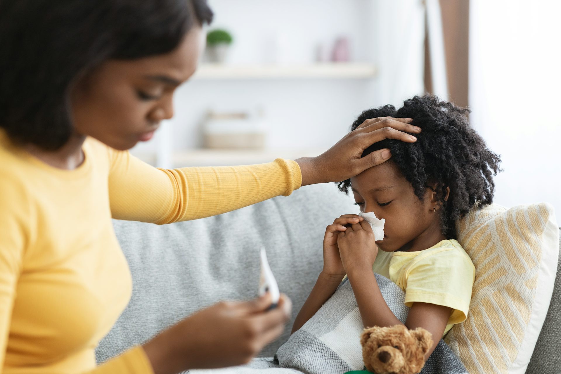 A mother checks her child's temperature with a thermometer while resting her hand on the child's head. The girl is blowing her nose with a tissue.