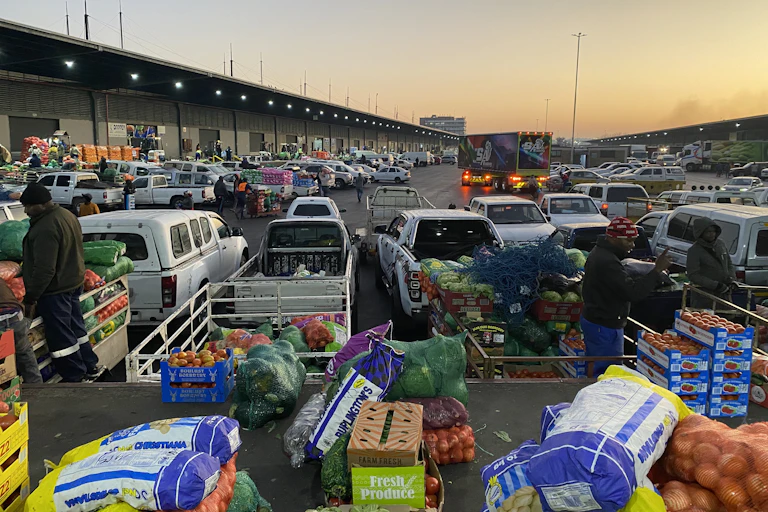 A bustling market with several cars loaded with crates of fruit and vegetables like tomatoes and apples.