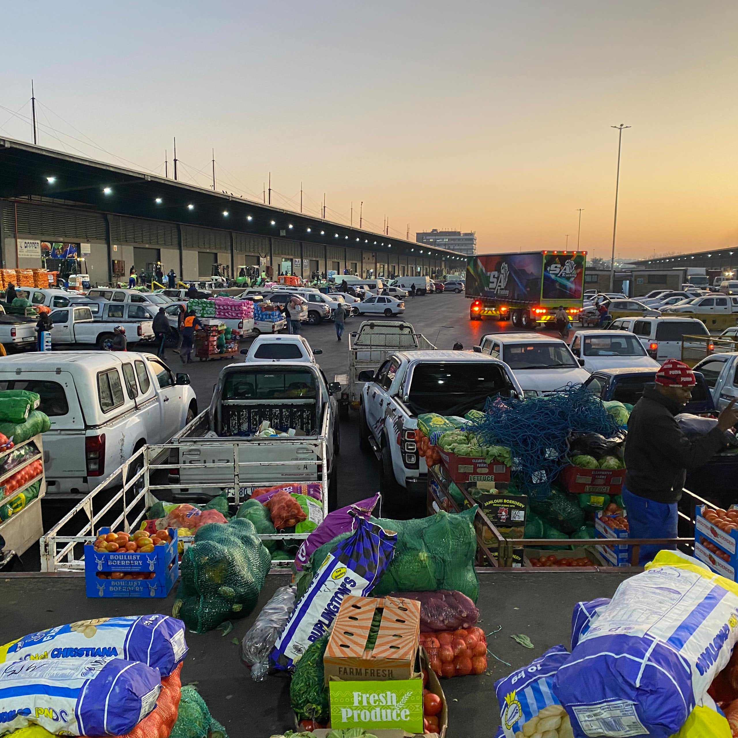 A bustling market with several cars loaded with crates of fruit and vegetables like tomatoes and apples.