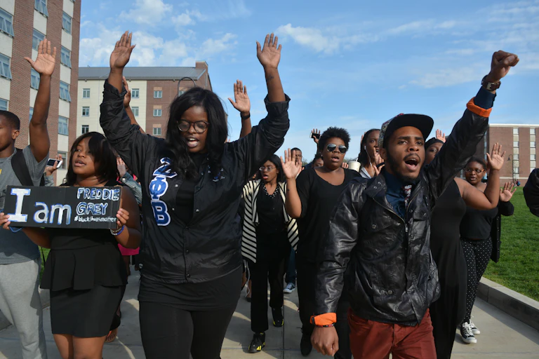 A group of young Black people walk together and some hold their hands up in the air. They walk past large buildings.