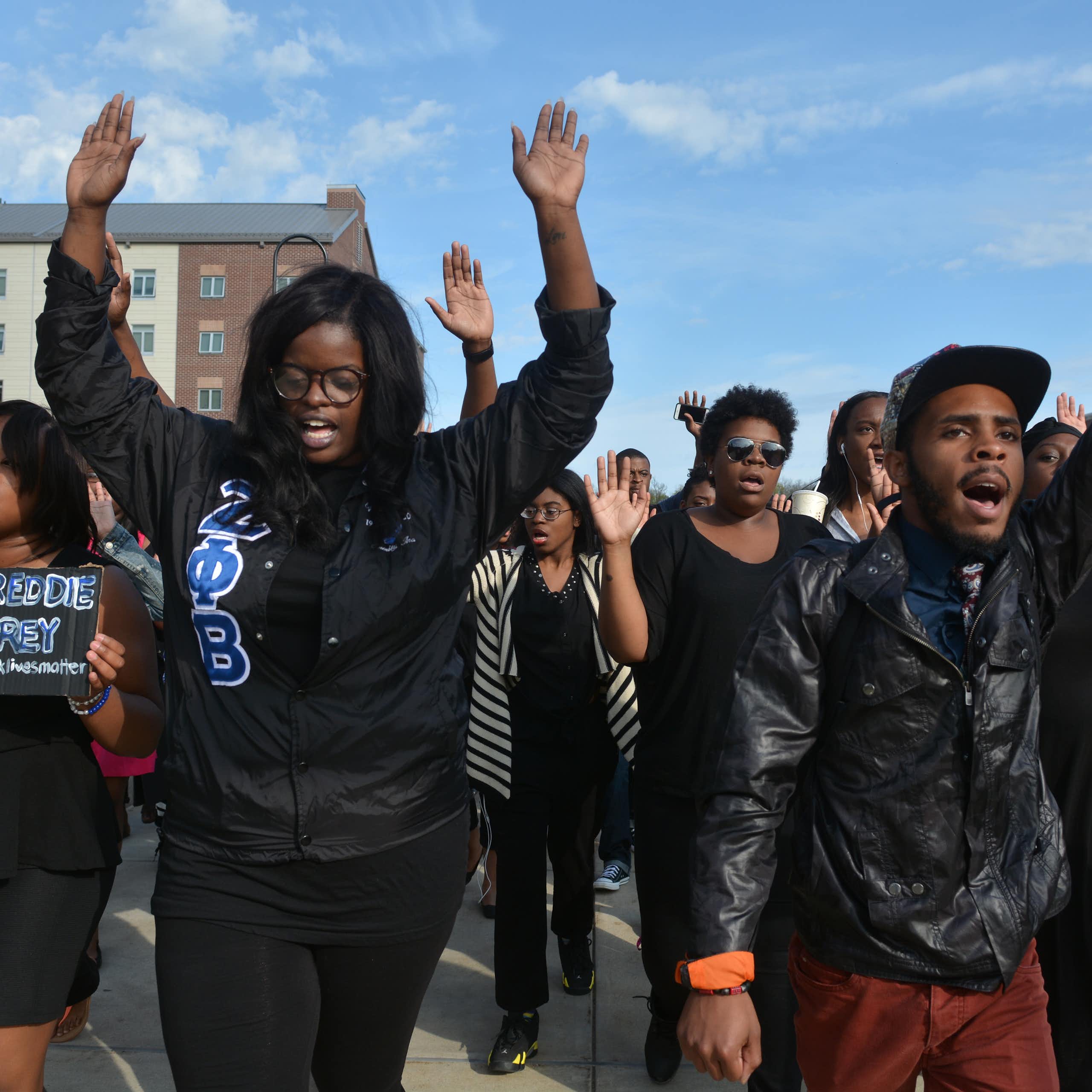 A group of young Black people walk together and some hold their hands up in the air. They walk past large buildings.