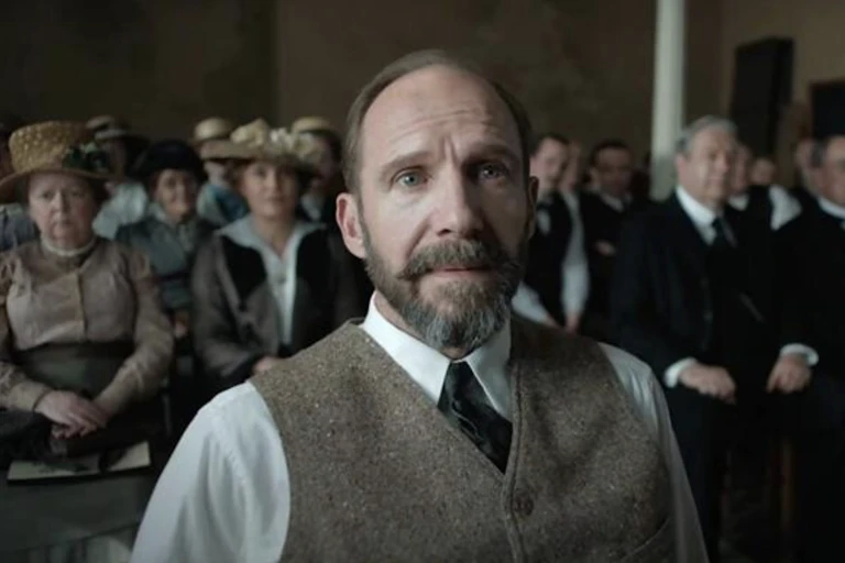 A pleasant-looking man from the Edwardian period standing in front in front of seated group of people.