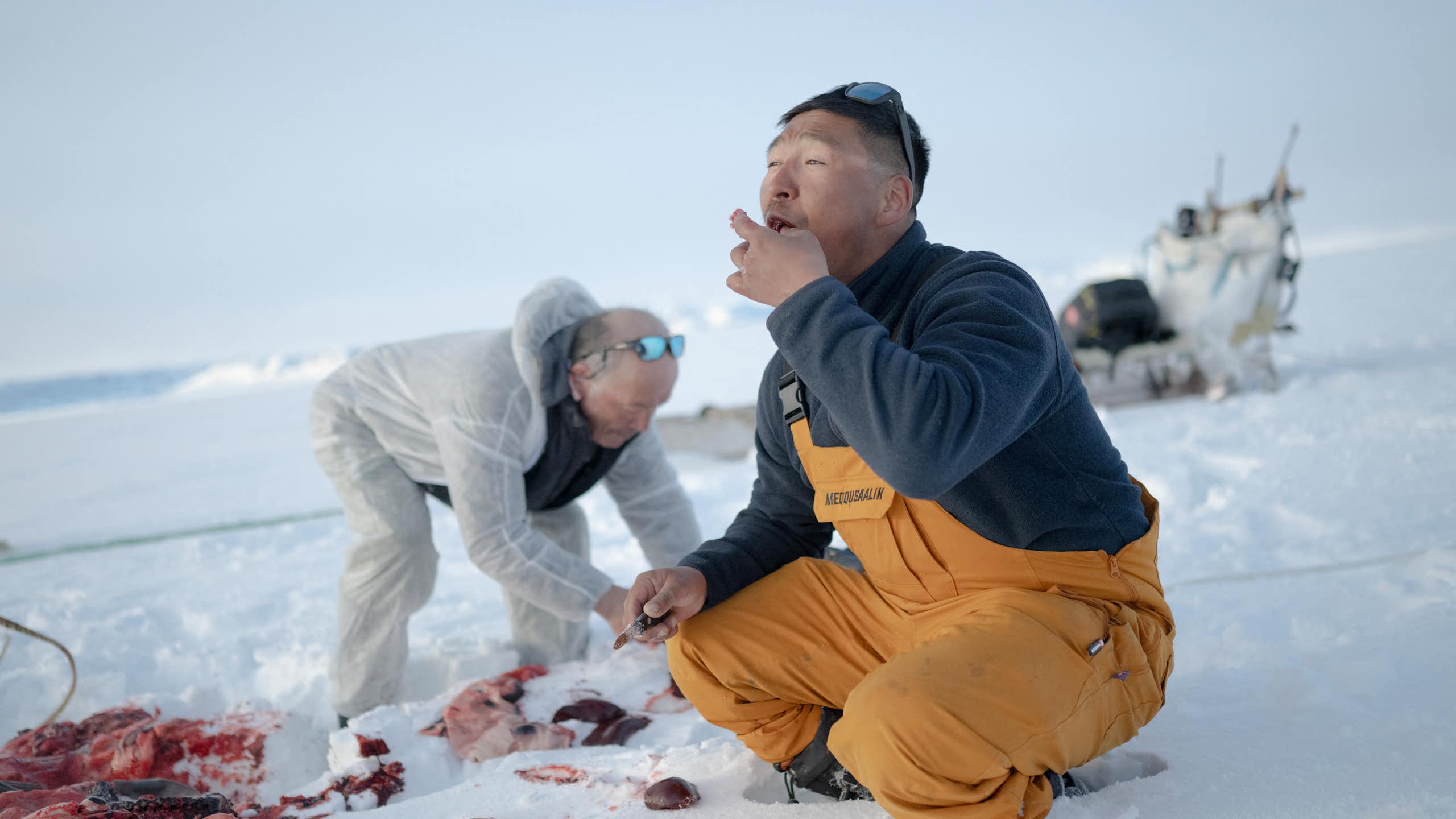 two men on a snow-covered surface near the bloody remains of an animal