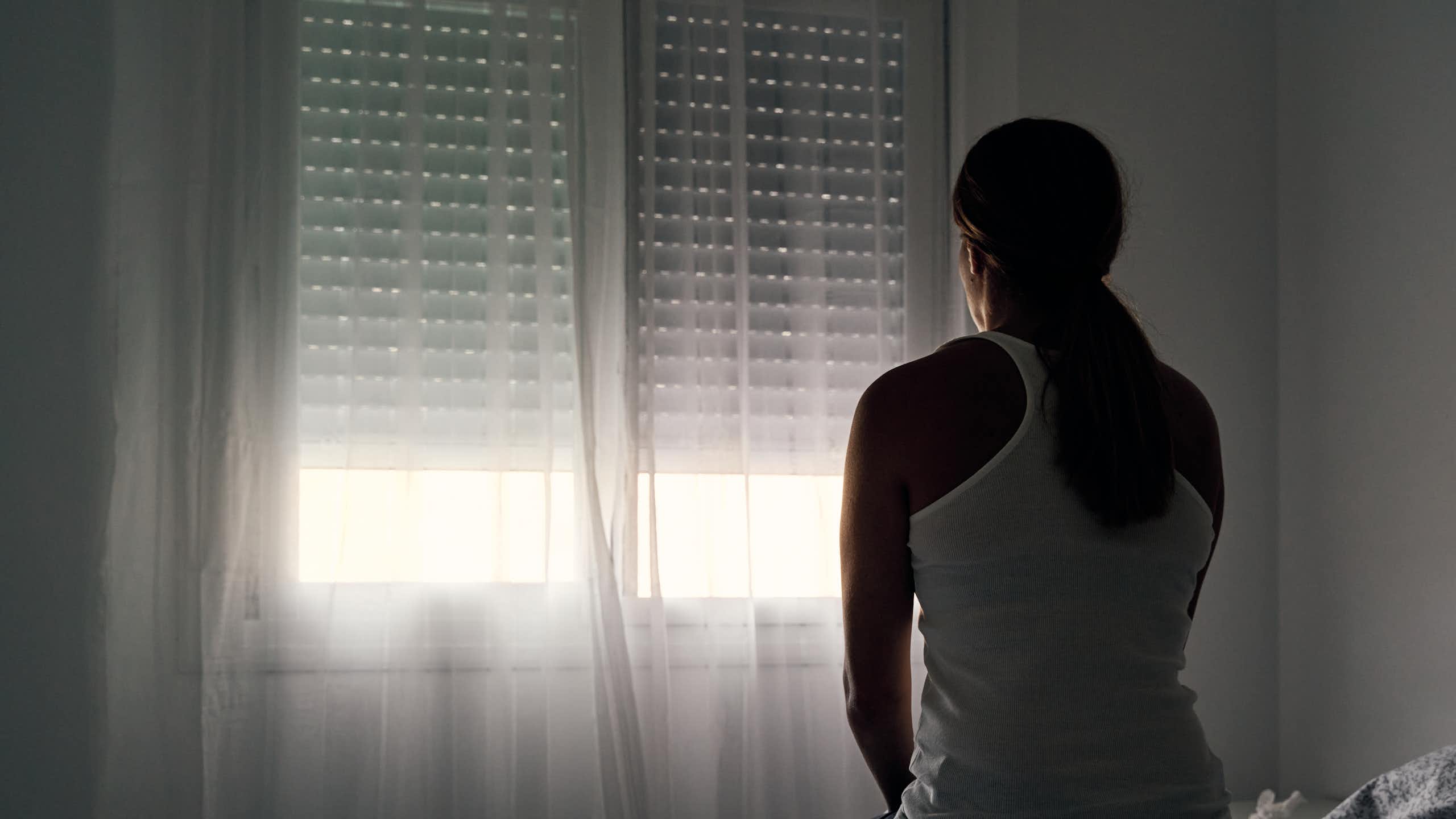 A woman sits in a dark room facing a window with cracked open shades.