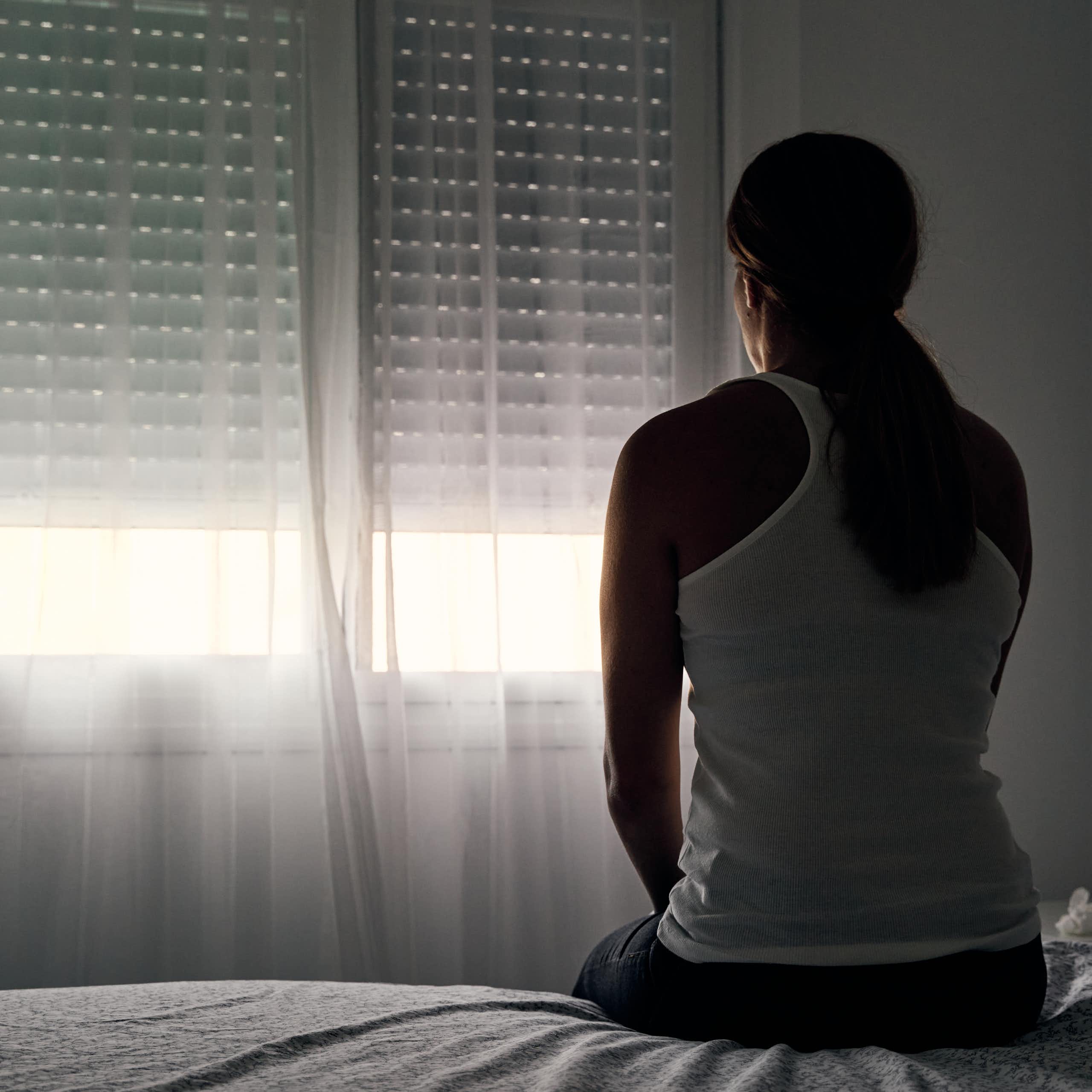 A woman sits in a dark room facing a window with cracked open shades.