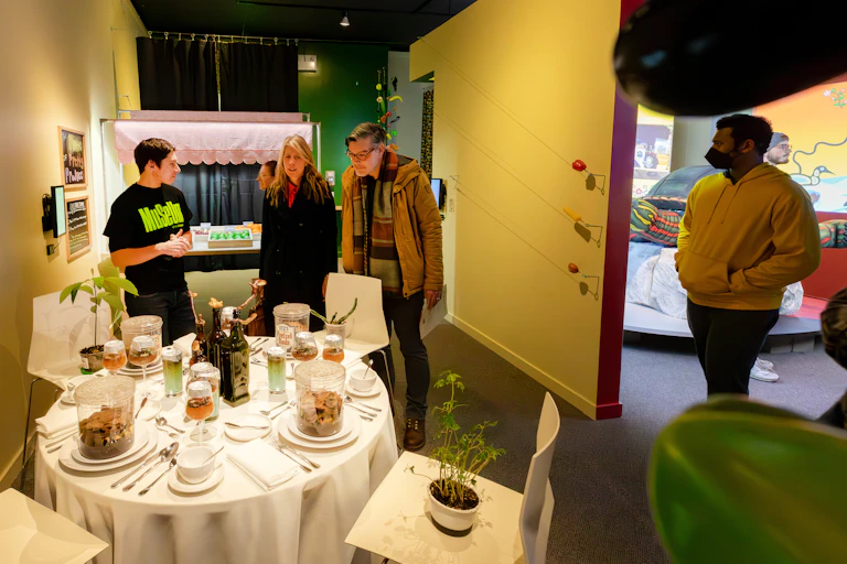 A couple speaks to a young man wearing a T-shirt that reads "MuSeUm" in bright green type. They are standing in front of a round table set for a meal. A plant sits in one of the chairs.