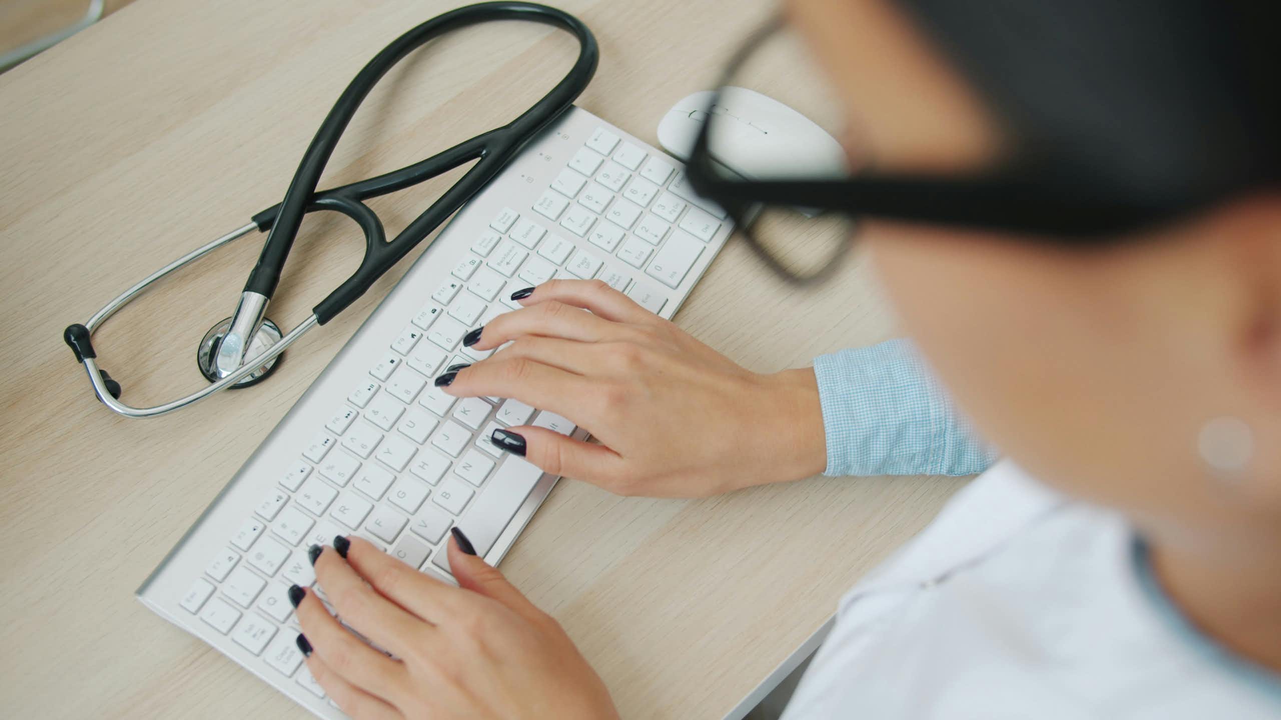 A person typing on a keyboard with a stethoscope on the desk