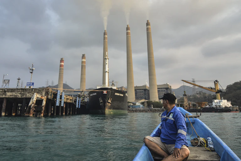 A man in a small boat turns to look at the five stacks of a coal-fired power plant along the shore behind him.