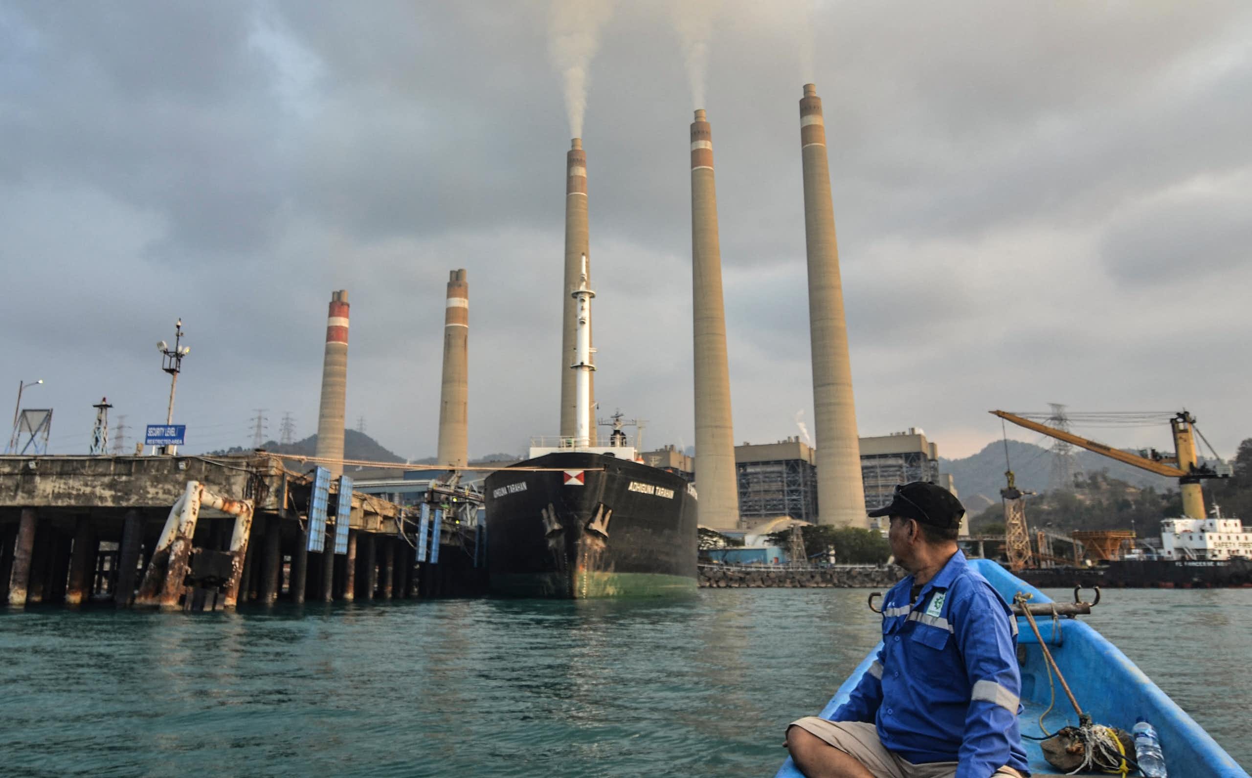 A man in a small boat turns to look at the five stacks of a coal-fired power plant along the shore behind him.