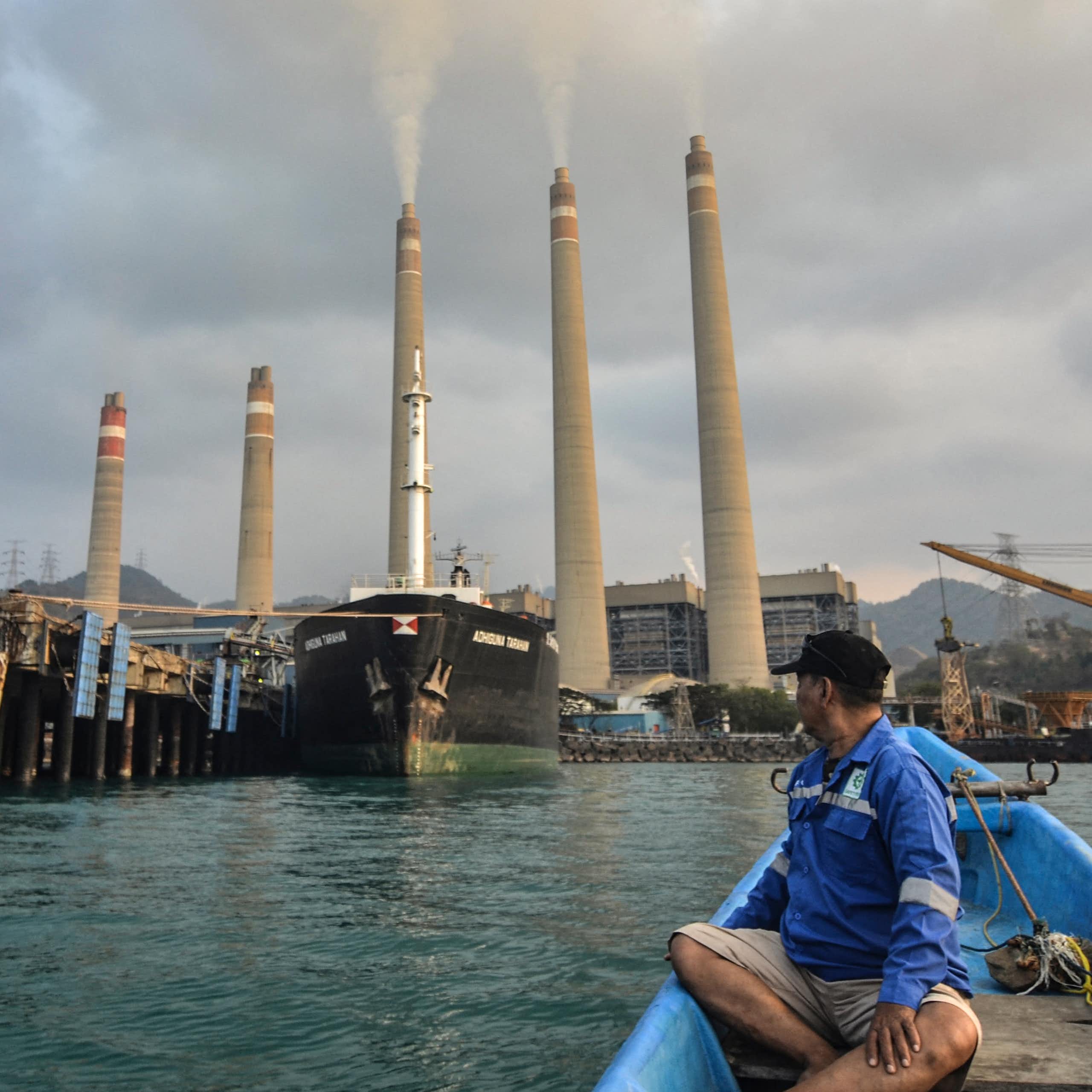 A man in a small boat turns to look at the five stacks of a coal-fired power plant along the shore behind him.